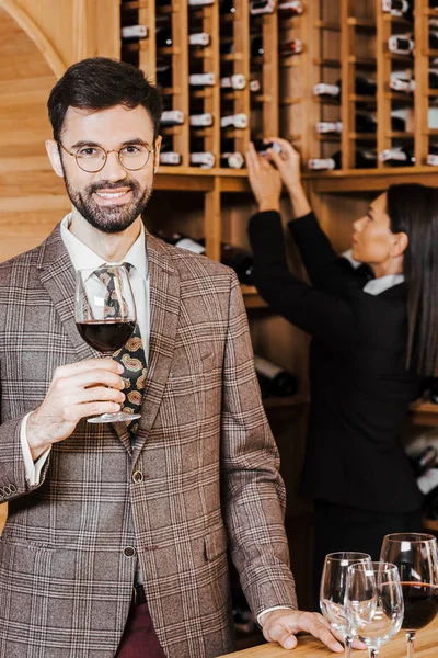 Steward di vino femminile che prende bottiglia dallo scaffale per il cliente mentre guarda la fotocamera in enoteca — Foto stock