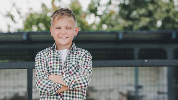 Niño feliz con camisa a cuadros de pie con los brazos cruzados y sonriendo a la cámara en la granja - foto de stock