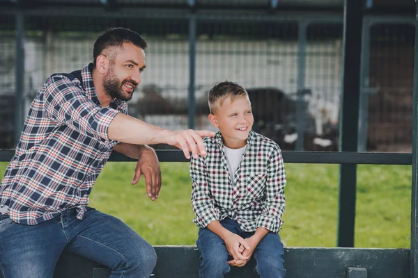 Happy father and son sitting together and looking away at ranch — Stock Photo