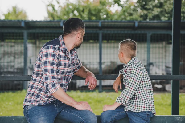 Side view of father and son in checkered shirts sitting together and looking away at ranch — Stock Photo
