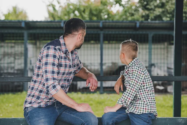 Vista laterale di padre e figlio in camicie a scacchi seduti insieme e guardando lontano ranch — Foto stock