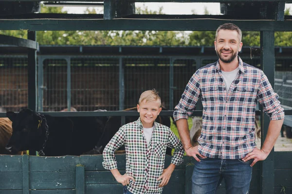 Heureux père et fils debout avec les mains sur la taille et souriant à la caméra à la ferme — Photo de stock