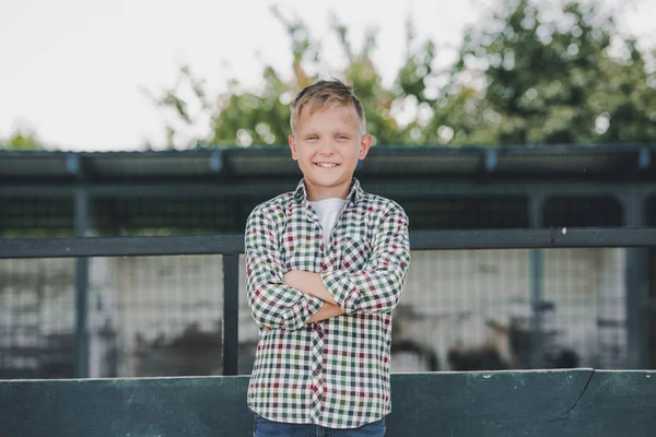 Adorabile bambino in camicia a scacchi in piedi con le braccia incrociate e sorridente alla fotocamera in fattoria — Foto stock
