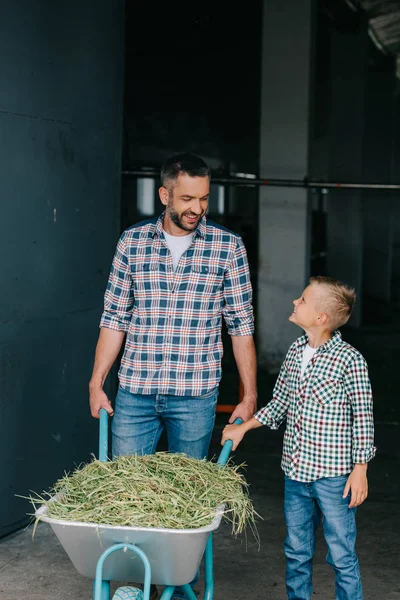 Happy father and son pushing wheelbarrow with grass and smiling each other in stall — Stock Photo