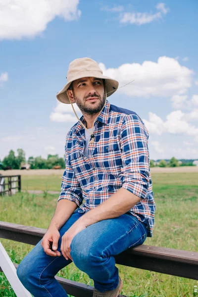 Pensativo agricultor de mediana edad en sombrero de panama sentado en la cerca y mirando hacia el campo - foto de stock