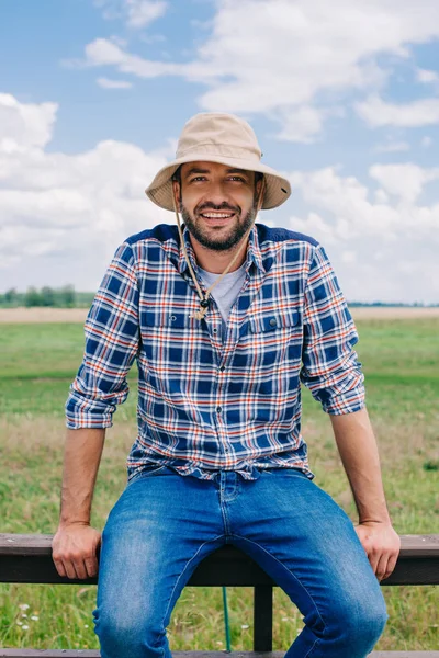 Handsome middle aged farmer in panama hat sitting on fence and smiling at camera — Stock Photo