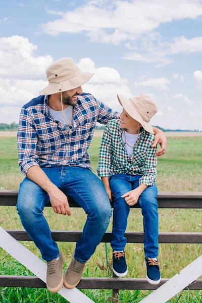 Feliz padre e hijo en sombreros panama sentados en la cerca y sonriendo unos a otros en la granja - foto de stock