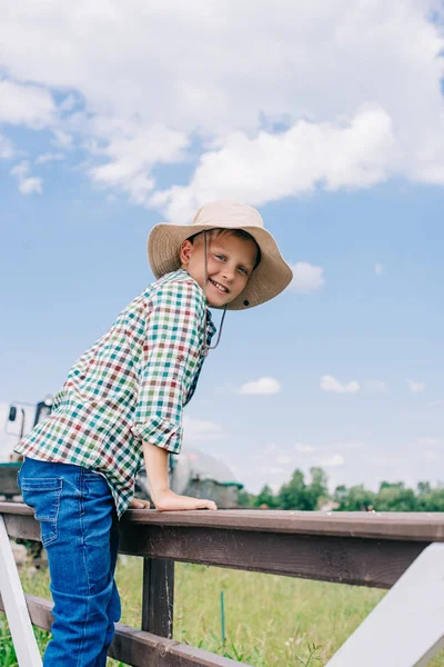 Cheerful child in panama hat climbing on fence and smiling at camera on farm — Stock Photo
