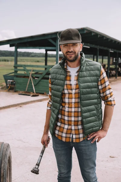 Handsome middle aged farmer in cap smiling at camera while standing with hand on waist at ranch — Stock Photo