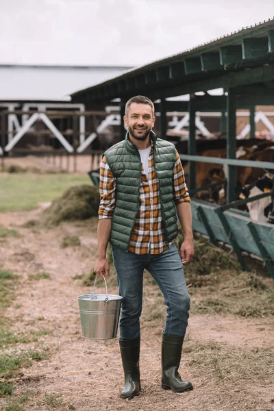 Guapo granjero macho sosteniendo cubo y sonriendo a cámara en establo - foto de stock