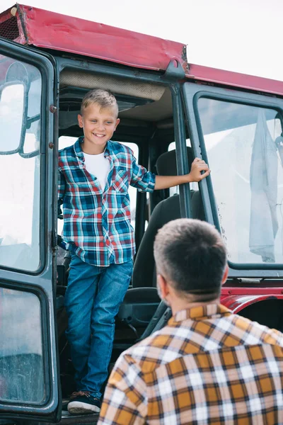 Vista trasera de padre mirando feliz hijo de pie en tractor - foto de stock