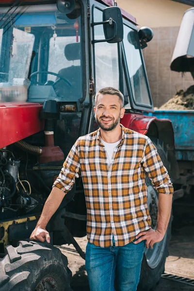 Hermoso agricultor de mediana edad de pie cerca del tractor y sonriendo a la cámara - foto de stock