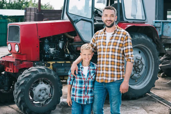 Feliz pai e filho em camisas quadriculadas sorrindo para a câmera enquanto estão juntos trator puro na fazenda — Fotografia de Stock