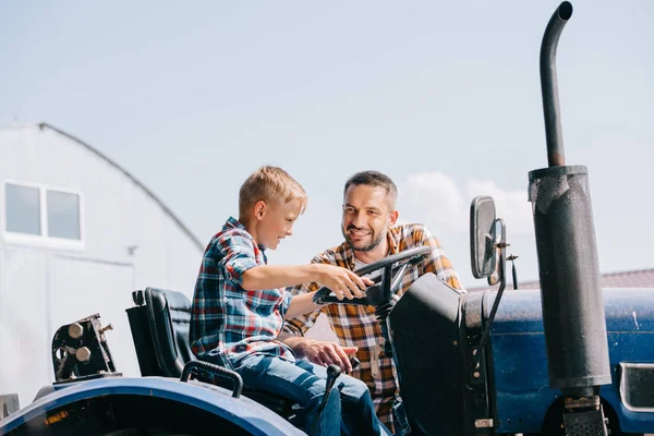 Feliz padre mirando sonriente hijo sentado en tractor en granja - foto de stock