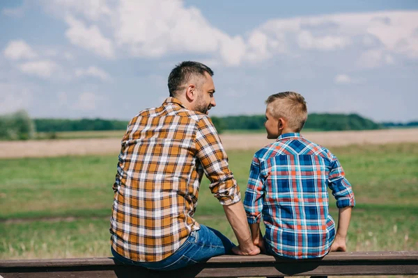 Rückansicht von Vater und Sohn, die auf dem Zaun sitzen und einander auf dem Bauernhof anlächeln — Stockfoto