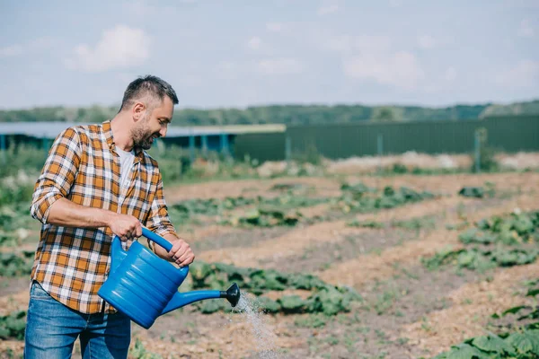 Красивый фермер в клетчатой рубашке держит лейку и работает в поле — стоковое фото