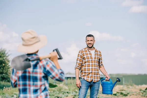 Селективный фокус мальчика с лопатой глядя на улыбающегося отца с поливной банкой на поле — стоковое фото