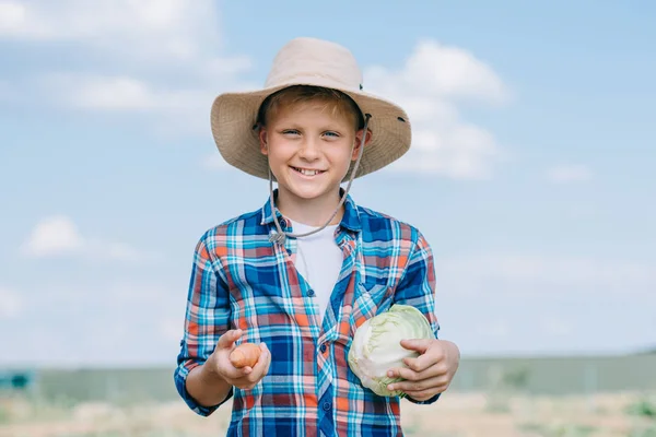 Happy boy in panama hat holding ripe vegetables and smiling at camera outdoors — Stock Photo