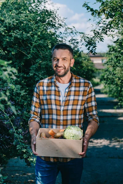 Happy farmer holding box with fresh vegetables and smiling at camera — Stock Photo