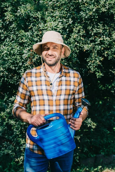 Handsome male farmer in checkered shirt holding watering can and smiling at camera outdoors — Stock Photo