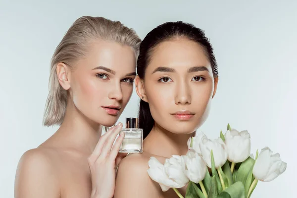 Attractive multicultural girls posing with tulips and bottle of perfume, isolated on grey — Stock Photo
