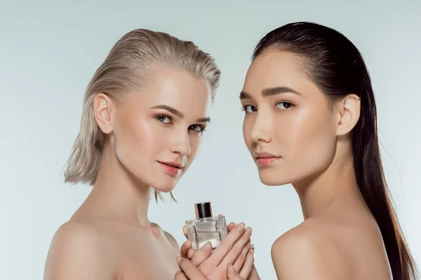 Naked multiethnic girls posing with bottle of perfume, isolated on grey — Stock Photo