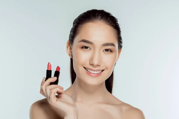 Приваблива усміхнена азіатська жінка тримає дві помади, ізольована на сірому — стокове фото