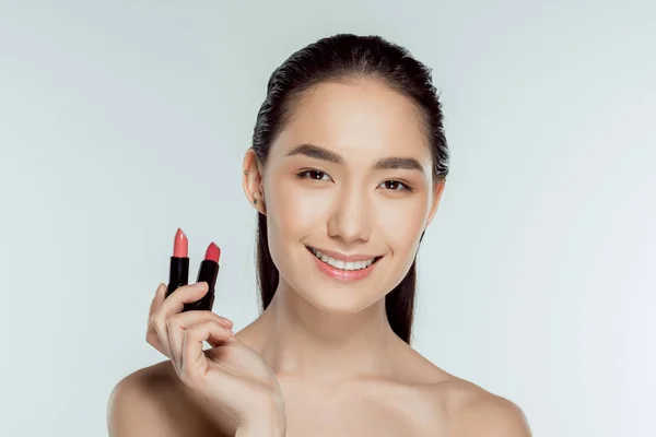 Attractive smiling asian woman holding two lipsticks, isolated on grey — Stock Photo