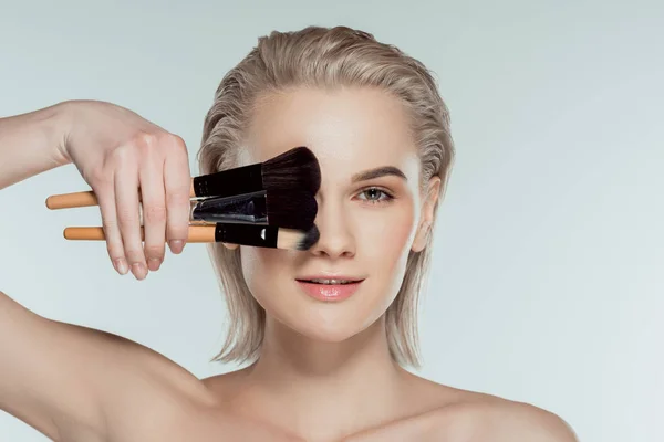 Femme blonde tenant des pinceaux cosmétiques, isolée sur gris — Photo de stock