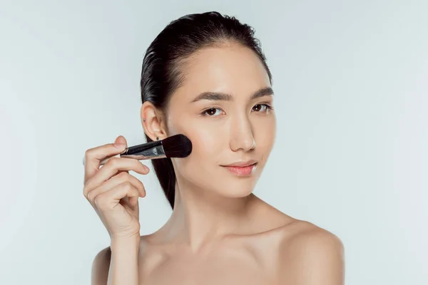 Menina asiática aplicando blushes com escova cosmética, isolado em cinza — Fotografia de Stock