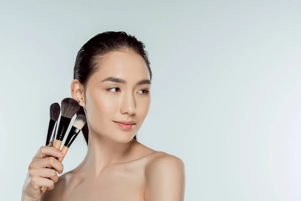Atractiva mujer asiática sosteniendo cepillos de maquillaje, aislado en gris - foto de stock