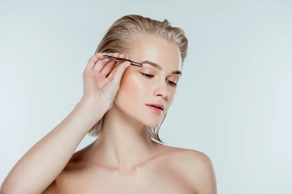 Beautiful woman correcting eyebrows with tweezers, isolated on grey — Stock Photo