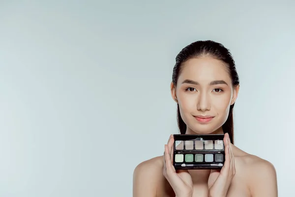 Atractiva chica asiática sosteniendo paleta con sombras de ojos, aislado en gris - foto de stock