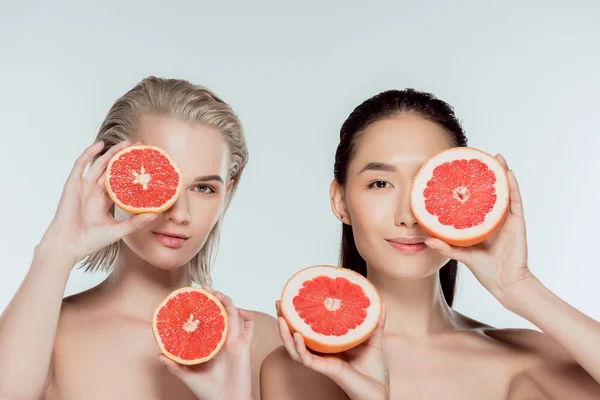 Красивые многоэтнические девушки, позирующие с половинками грейпфрута, изолированные на сером, концепция ухода за кожей — стоковое фото