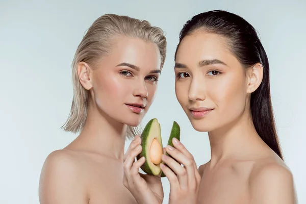 Niñas multiculturales posando con dos mitades de aguacate, aisladas en gris, concepto de cuidado de la piel - foto de stock