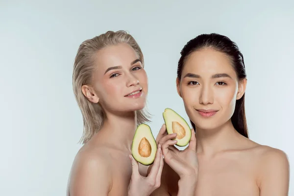 Chicas desnudas multiétnicas posando con dos mitades de aguacate, aisladas en gris, concepto de cuidado de la piel - foto de stock