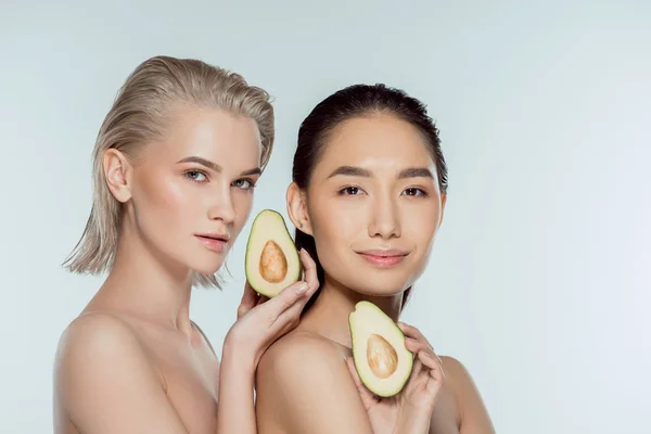 Niñas puras multiétnicas posando con dos mitades de aguacate, aisladas en gris, concepto de cuidado de la piel - foto de stock