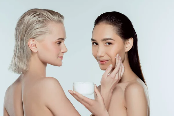 Meninas nuas multiétnicas aplicando creme facial, isolado em cinza, conceito de cuidados com a pele — Fotografia de Stock