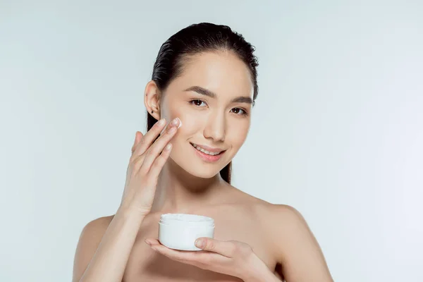 Atractiva mujer asiática aplicando crema facial, aislado en gris - foto de stock