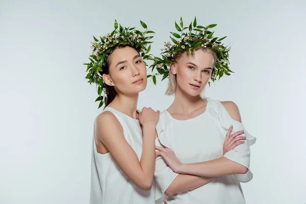 Attraktive multiethnische Mädchen posieren in weißen Kleidern und floralen Kränzen, isoliert auf grau, natürliche Schönheit — Stockfoto
