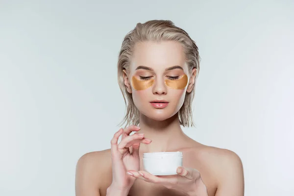Femme avec des patchs pour les yeux et crème pour le visage dans un récipient en plastique, isolé sur gris — Photo de stock