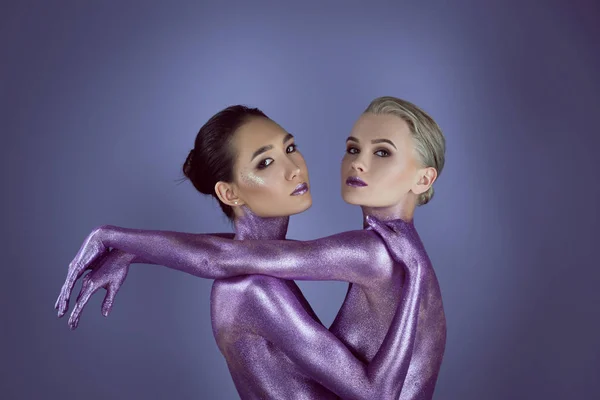 Мультикультурные привлекательные девушки в ультрафиолетовом блеске обнимая друг друга, изолированные на фиолетовый — стоковое фото