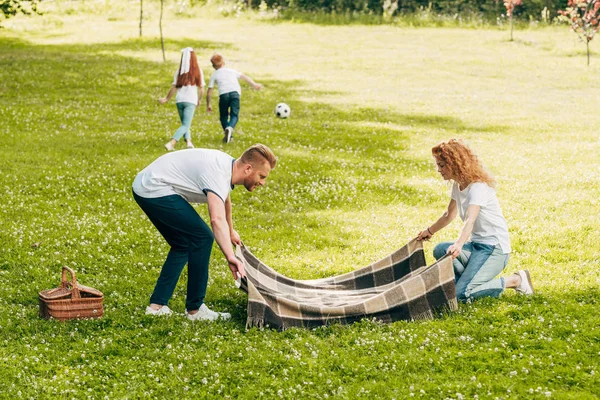 Padres sosteniendo cuadros mientras los niños juegan con la pelota en el picnic en el parque - foto de stock