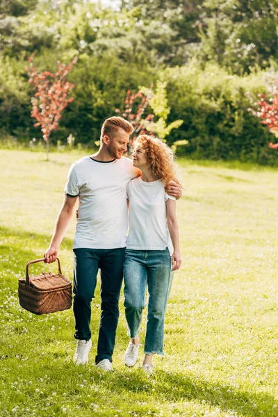 Счастливая молодая пара с корзинкой для пикника прогуливаясь по газону в парке — стоковое фото