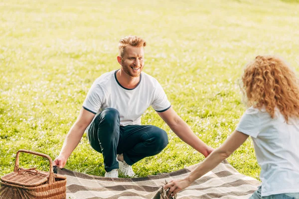 Happy redhead couple holding plaid and looking at each other at picnic in park — Stock Photo