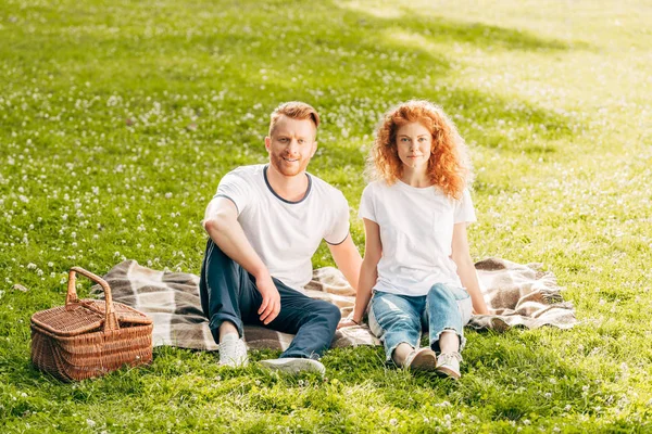 Glückliches Paar lächelt in die Kamera, während es zusammen auf Plaid beim Picknick im Park sitzt — Stockfoto