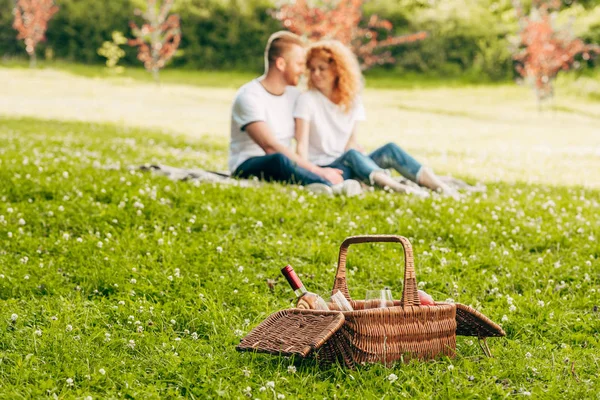Vista de cerca de la cesta de picnic con botella de vino y pareja sentada en el césped detrás en el parque - foto de stock