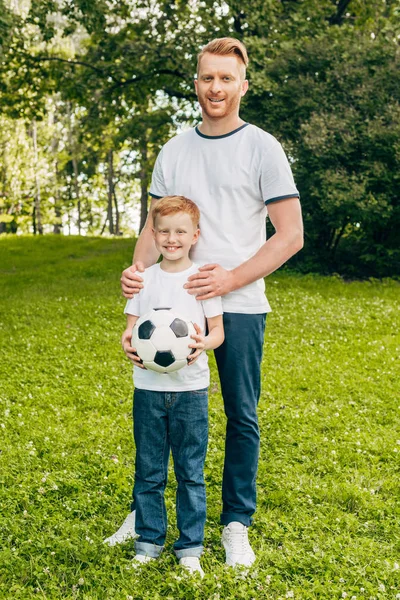 Heureux père et fils avec ballon de football debout ensemble et souriant à la caméra dans le parc — Photo de stock