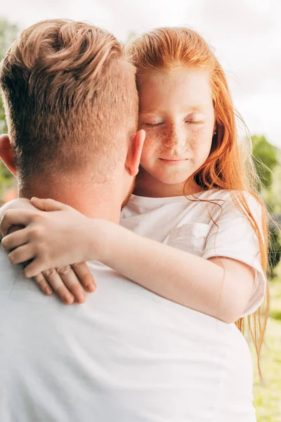 Adorable pelirroja niño con los ojos cerrados abrazando padre en parque - foto de stock
