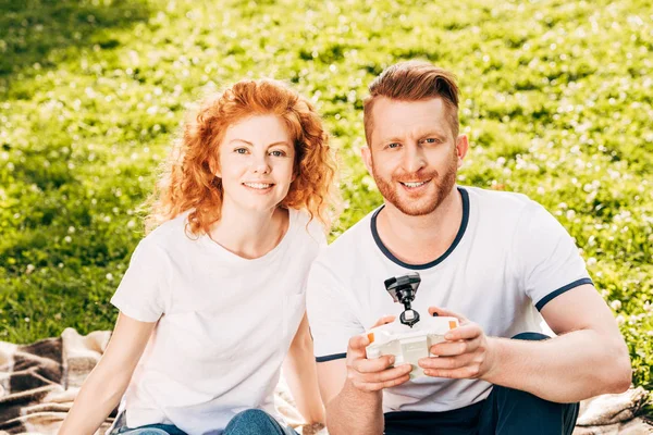 Щаслива пара грає з безпілотником і посміхається на камеру, сидячи на площині в парку — стокове фото